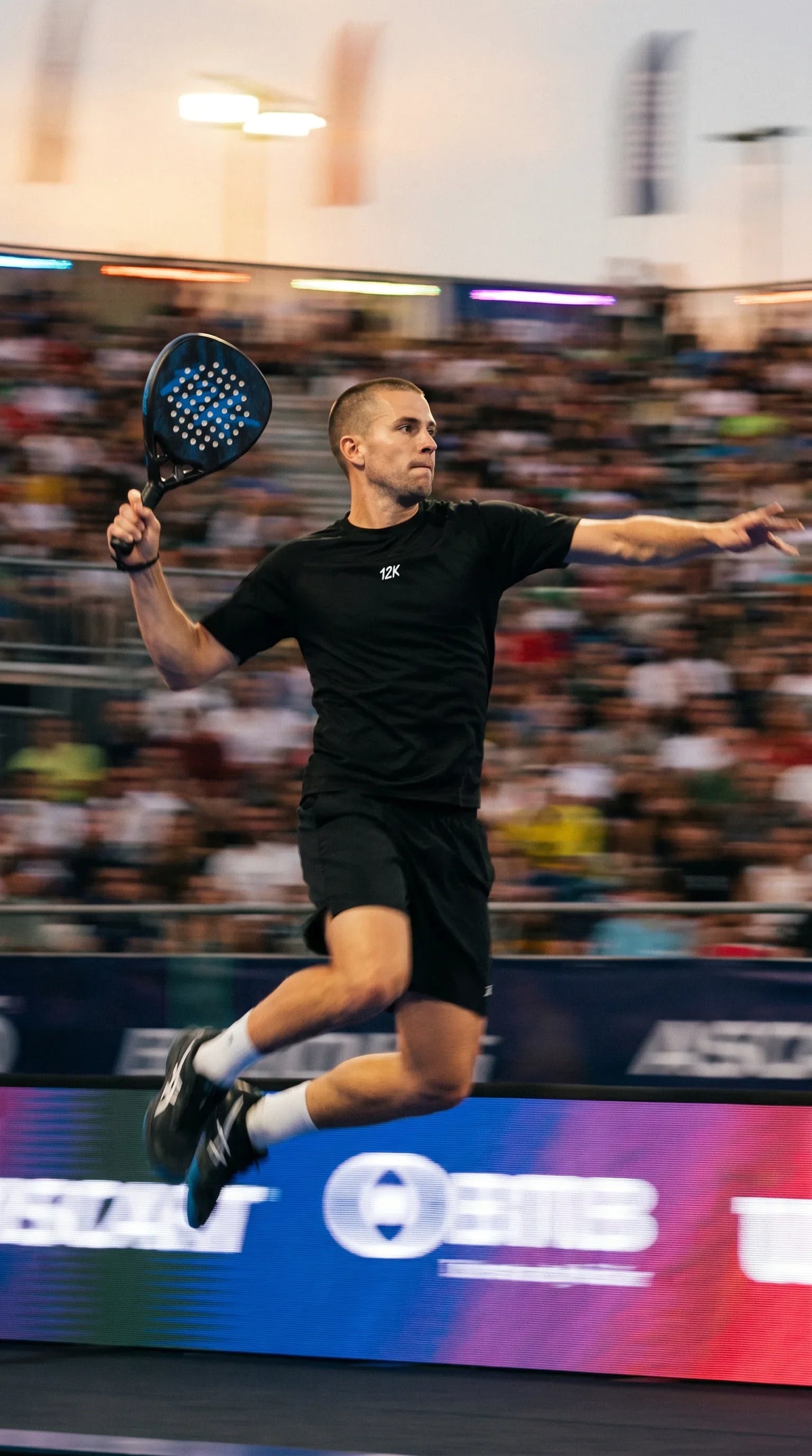12K Padel Shark Blue racket, player leaping for a smash in a stadium