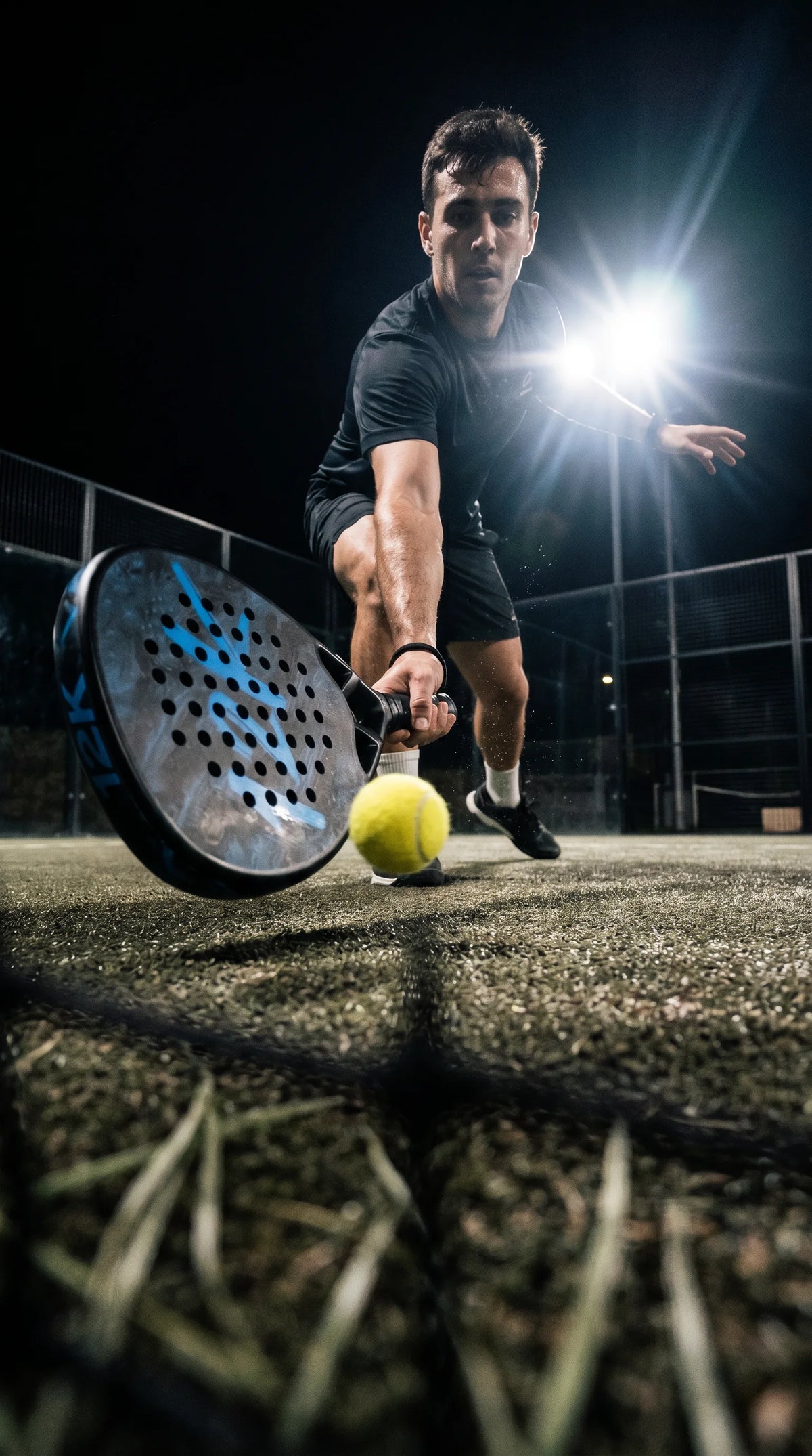 12k Padel Shark Blue racket, player digging out a low ball under floodlights