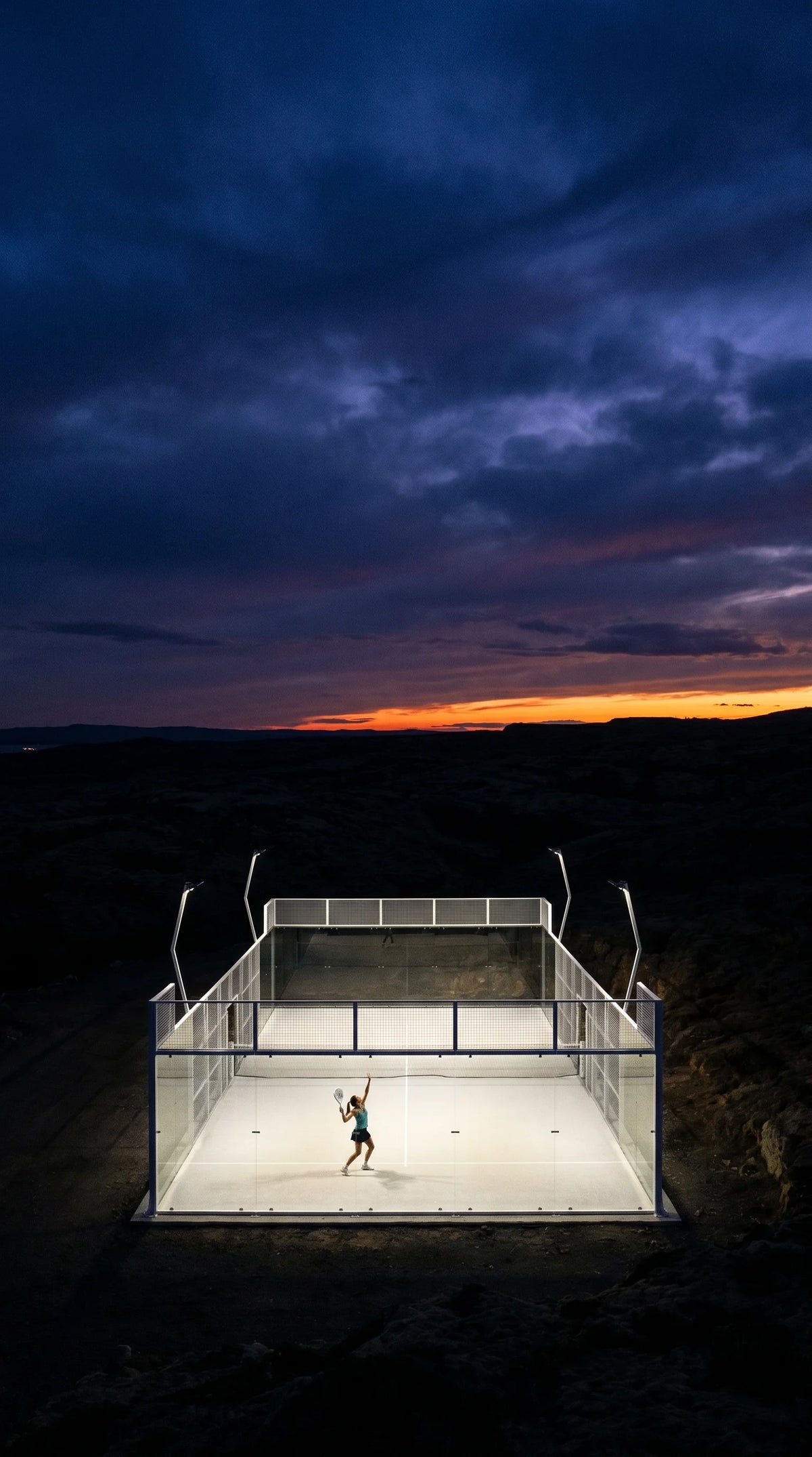 Floodlit glass padel court at dusk with a player serving