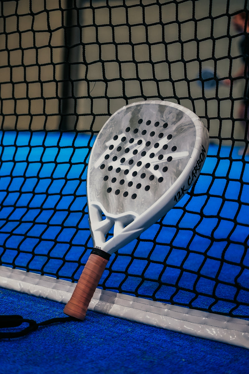 12K Padel Ice White racket with a peach overgrip resting against the net on a blue court