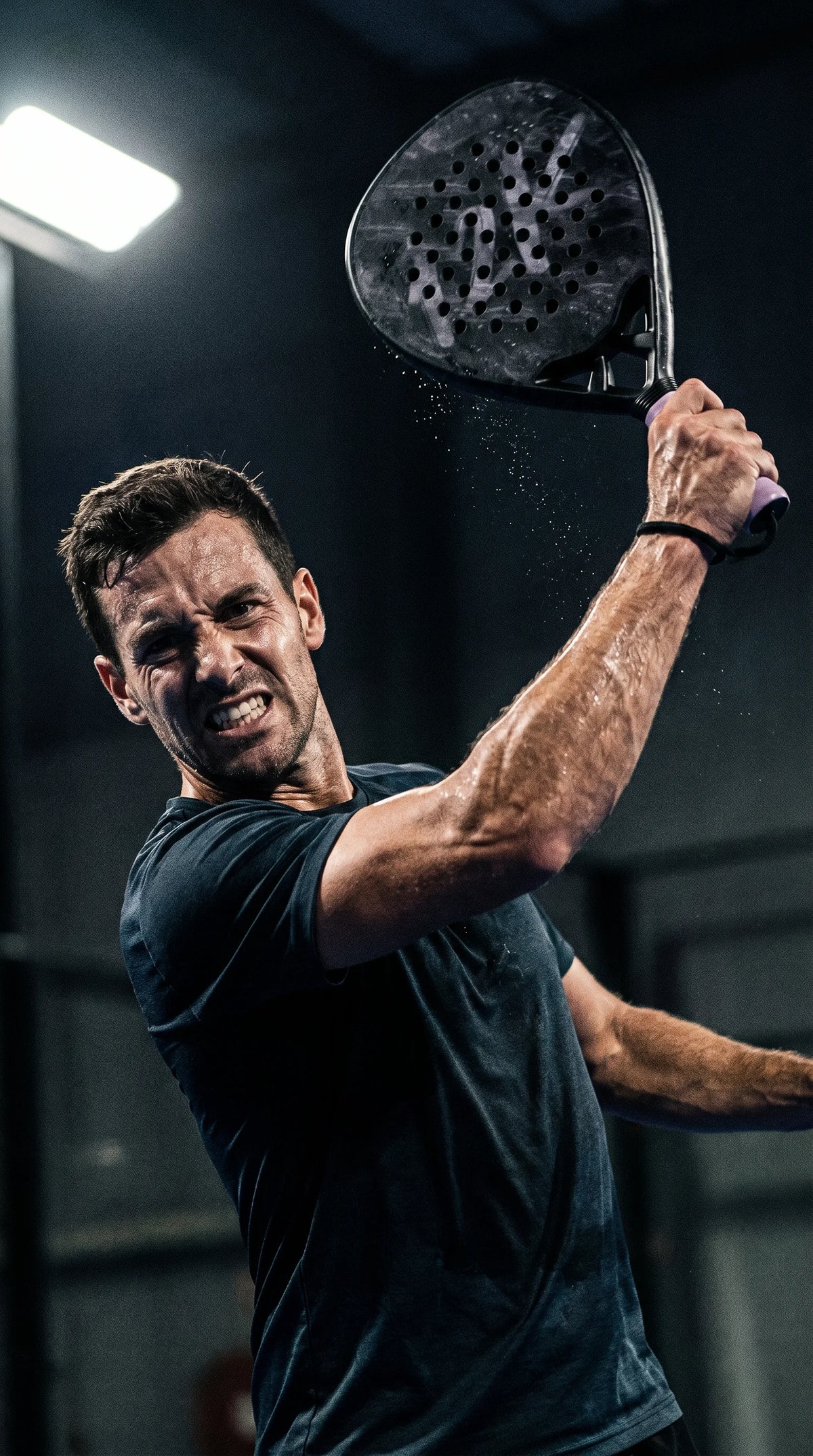 12k Padel Midnight Black racket, player winding up an overhead smash under floodlights